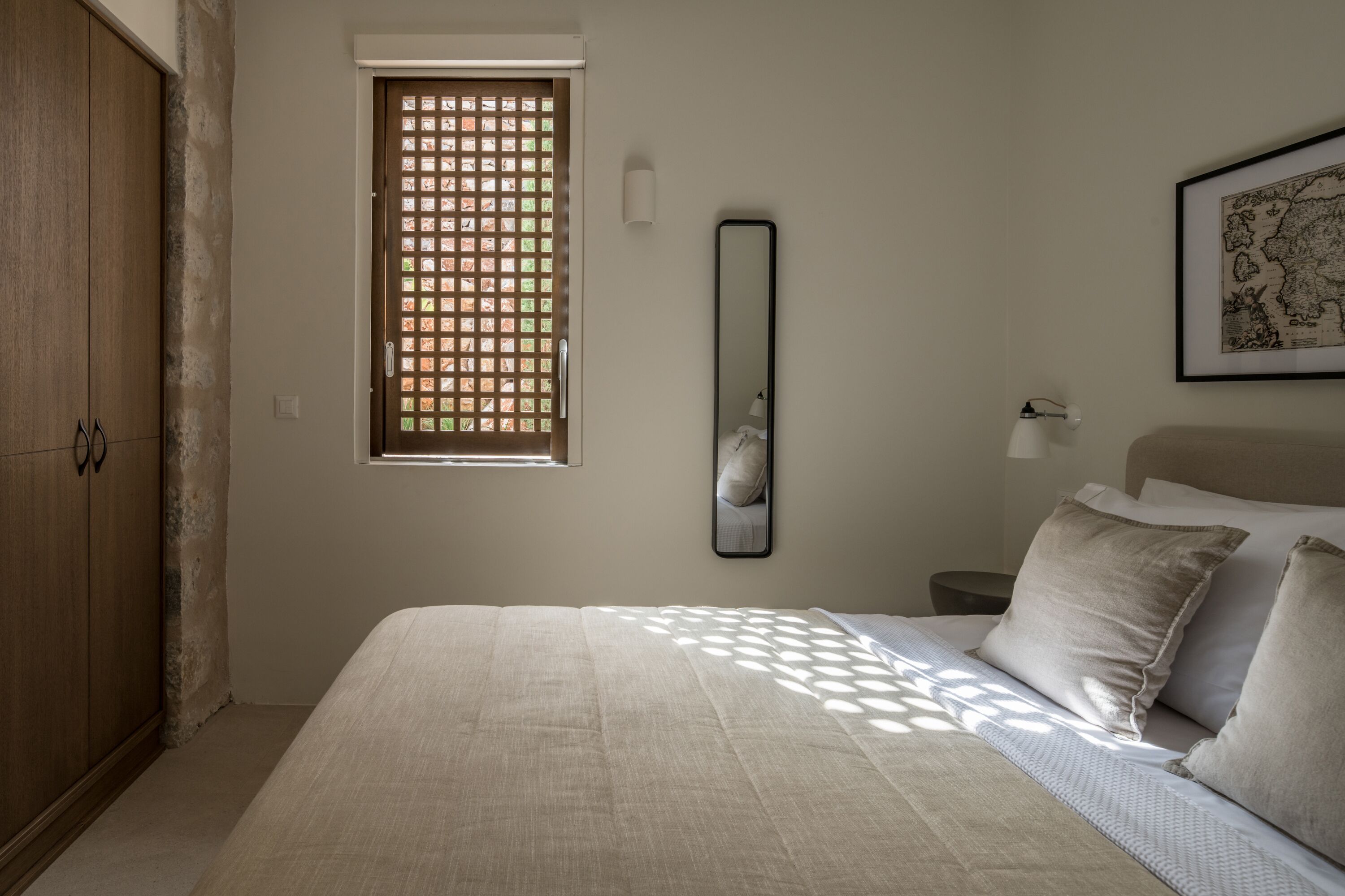 Un dormitorio con cama, ventana de diseño calado, mesita de noche con lámpara y un mapa en la pared.