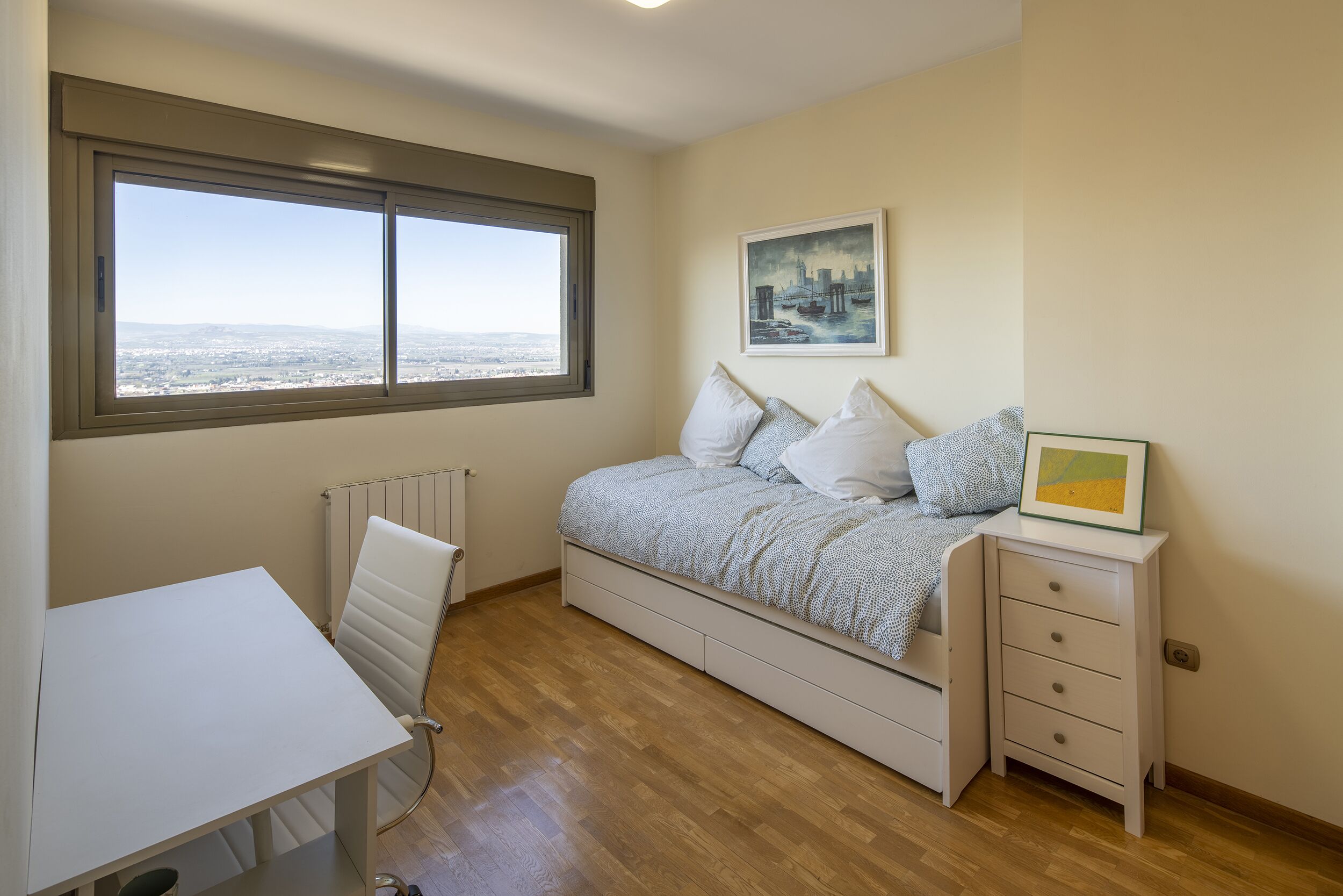 Une chambre à coucher comprenant un lit, un bureau, une chaise, un radiateur et une fenêtre donnant sur la ville.