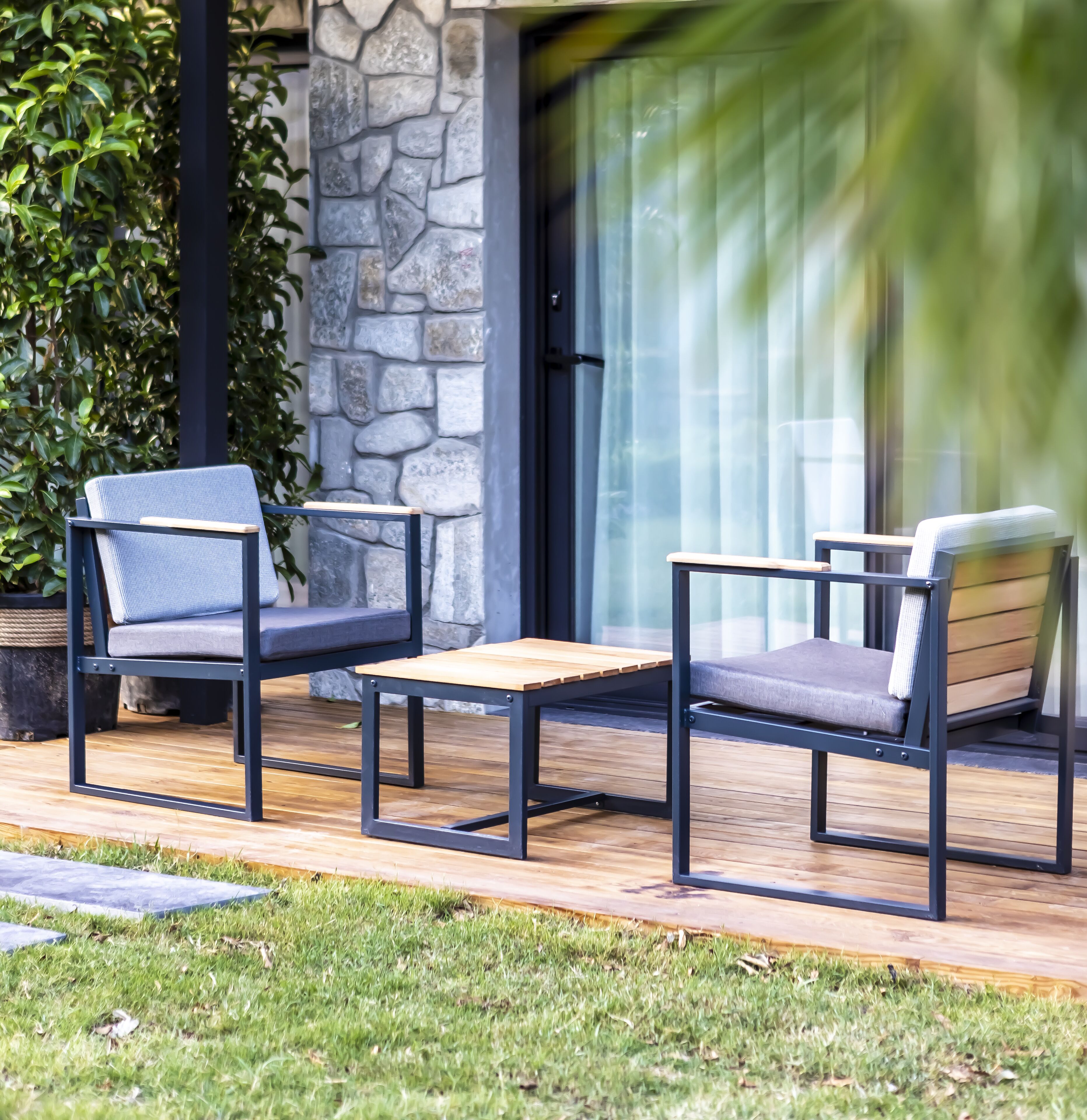 Two outdoor seating areas with metal frames and cushioned seats, positioned on a wooden deck with a stone wall and greenery in the background.