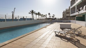 Piscina com espreguiçadeiras, um deck revestido e uma cerca com palmeiras ao fundo.