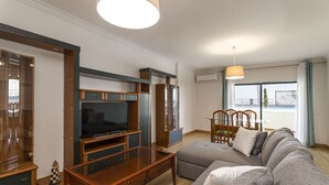 Uma sala de estar moderna com uma grande TV de tela plana, móvel para TV em madeira, sofá cinza e uma área de jantar com mesa e cadeiras.