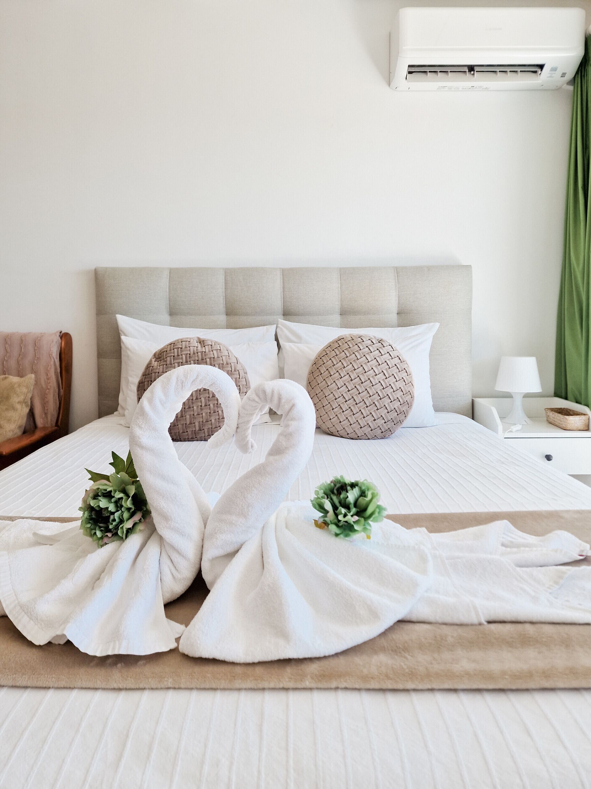 A neatly made bed with a swan-shaped towel arrangement, a headboard, and a bedside table with a lamp and a small basket.