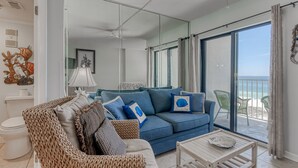 Ein modernes Wohnzimmer mit einem blauen Sofa, einem gewebten Sessel und einem Holztisch. Ein großer Spiegel spiegelt den Raum wider, und durch die Glastüren ist ein Balkon mit Meerblick zu sehen.