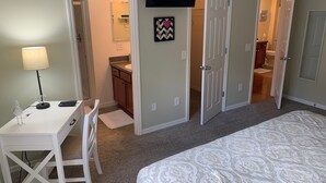 Un dormitorio con una cama, un escritorio con lámpara, un televisor montado en la pared y un baño visible a través de una puerta abierta.