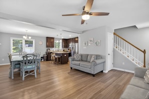 A spacious living area with a dining table, a sofa, a kitchen in the background, and a staircase.