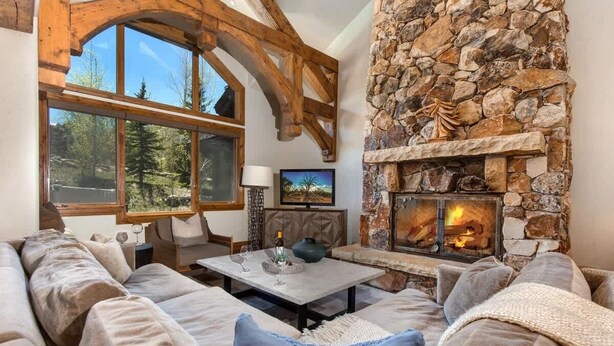 A cozy living room with a stone fireplace, wooden beams, and a large window overlooking a forest.