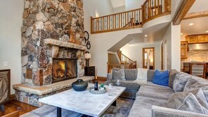 A spacious living area with a stone fireplace, a wooden staircase, and a kitchen in the background.