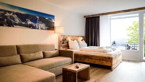 Une chambre d’hôtel moderne avec un lit en bois, un canapé, une petite table en bois et une grande fenêtre donnant sur l’extérieur.