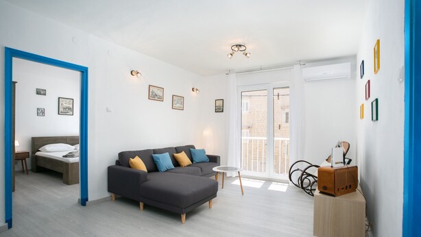 Ett modernt vardagsrum med en grå hörnsoffa, ett litet runt bord och en säng med ett träfärgat sänggavel.