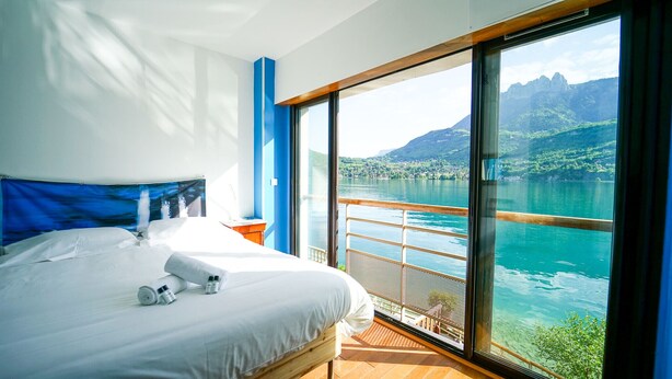 A modern hotel room with a large bed, a nightstand, and a view of a lake and mountains through a large window. - Feet in the water! (Duingt)
