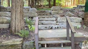 Una escalera de madera que sube hacia un muro de piedra, rodeada de árboles y follaje.