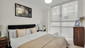A neatly arranged hotel room with a large bed, bedside lamps, a bedside table, a mirror, and a window with blinds. - One Bed Serviced Apt Moorgate (London)