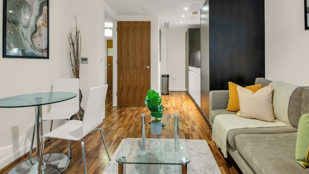 A modern living room with a glass coffee table, white chairs, a grey sofa with cushions, a wooden door, and a framed abstract painting on the wall. - One Bed Serviced Apt Moorgate (London)