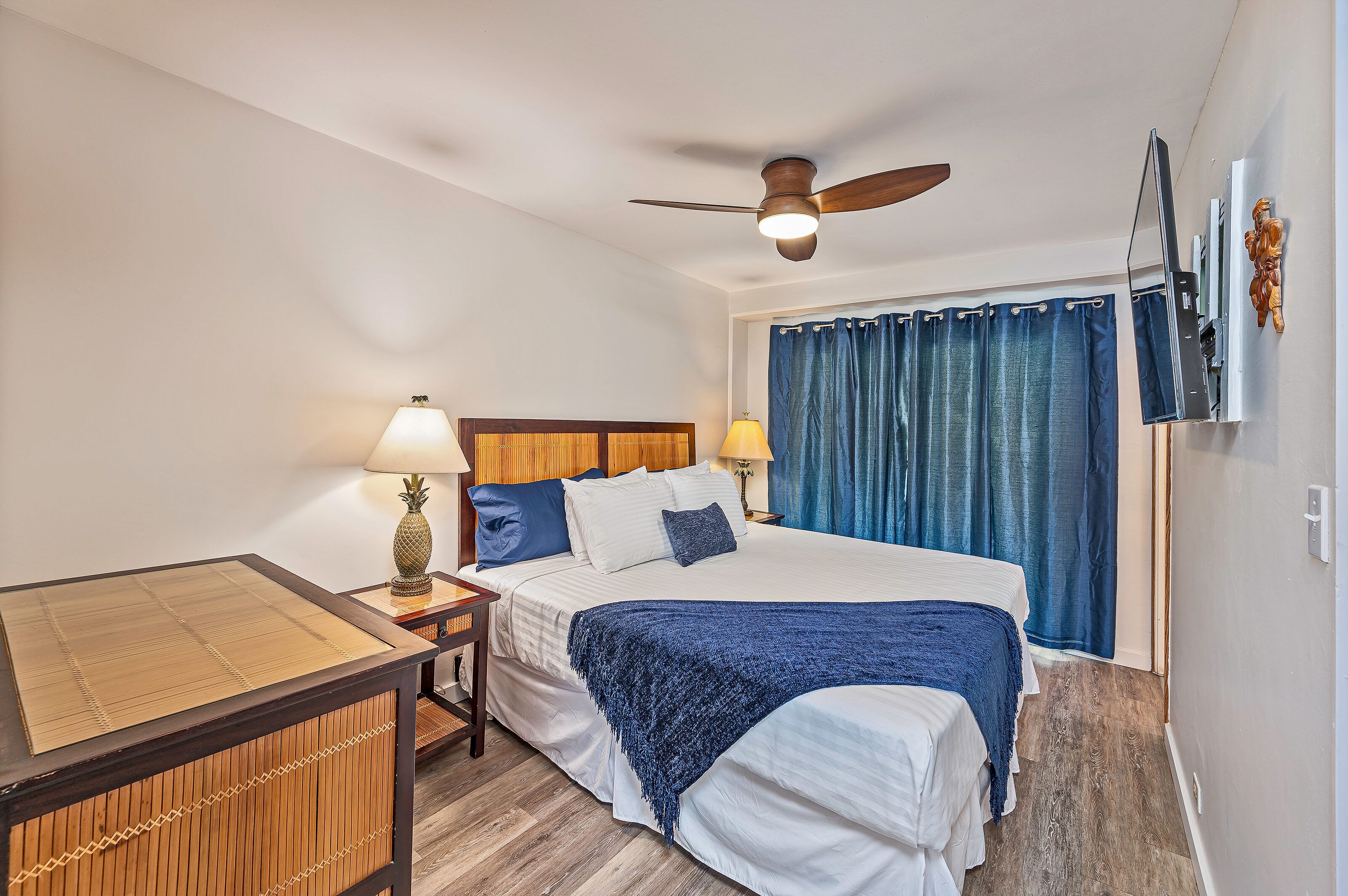 Quarto de hotel com cama, escrivaninha, ventoinha no teto e janela com cortinas.