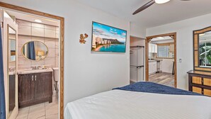 Ein Schlafzimmer mit Bett, Deckenventilator, Badezimmer mit Waschbecken, Spiegel und Toilette sowie Blick auf eine Küche und Essbereich.