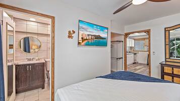 Une chambre avec un lit, un ventilateur de plafond, une salle de bain avec un lavabo, un miroir et des toilettes, et une vue sur la cuisine et la salle à manger.