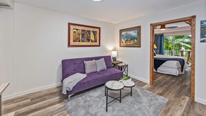 Ein modernes Wohnzimmer mit einem violetten Sofa, einem kleinen Tisch und einem Bett im Hintergrund. An den Wänden befinden sich gerahmte Bilder und durch ein Fenster ist ein Blick ins Freie möglich.