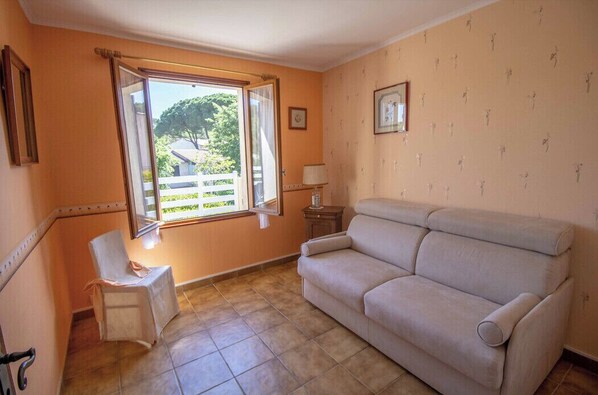 Une pièce avec un canapé beige, une petite table avec une lampe, une chaise et une fenêtre avec des rideaux.