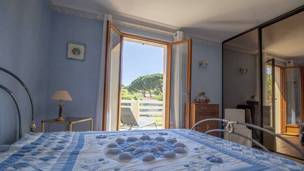 A bedroom with a large bed, a nightstand, a lamp, and a view through an open door to the outside. - Large Spacious House In Fréjus (Fréjus)