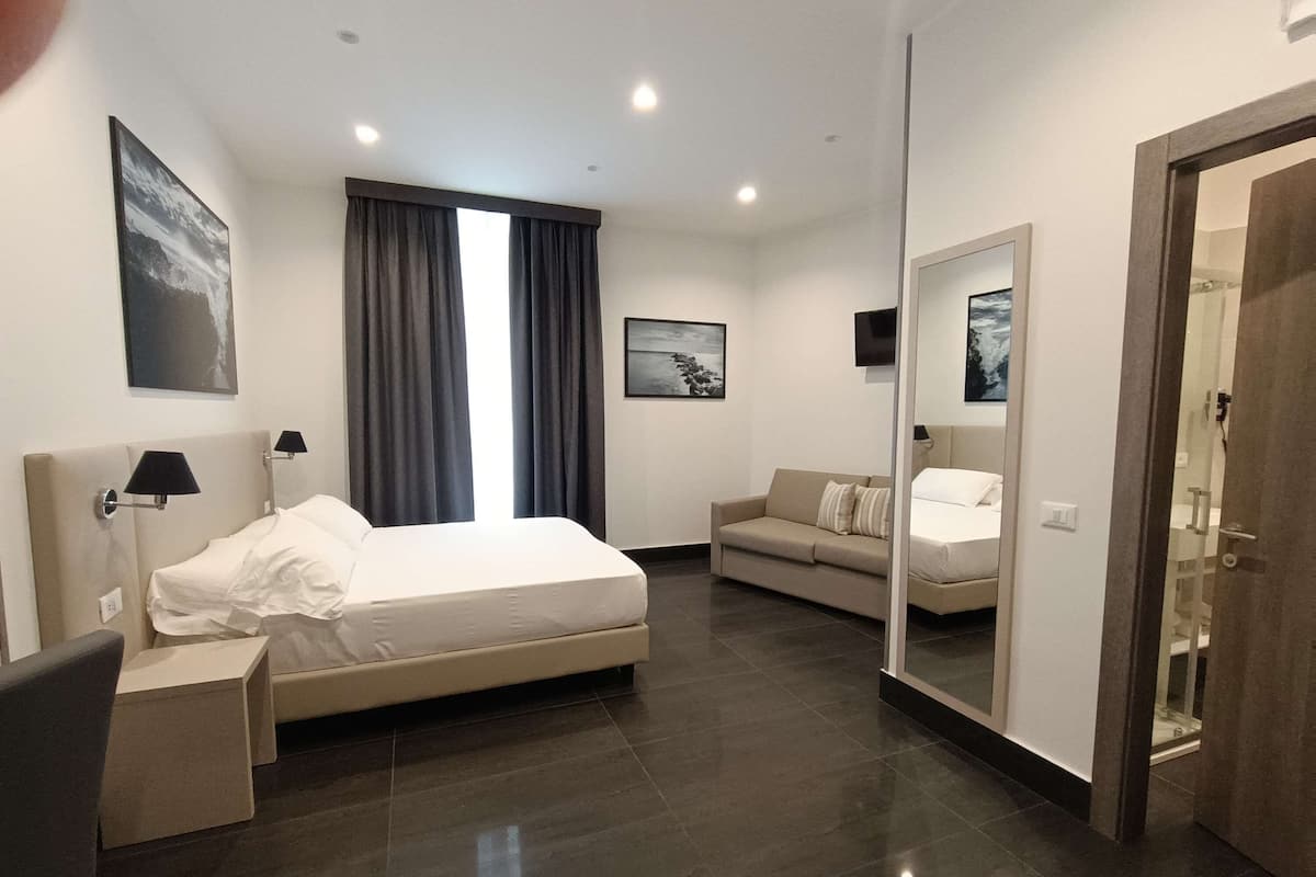 A modern hotel room with a large bed, a sofa, a desk, and a mirror.