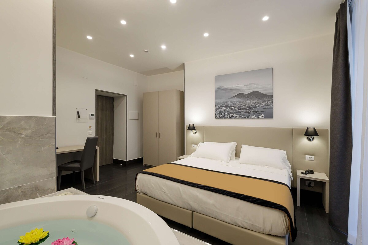 A modern hotel room with a large bed, a desk, a chair, a desk lamp, a nightstand, and a wall-mounted lamp.
