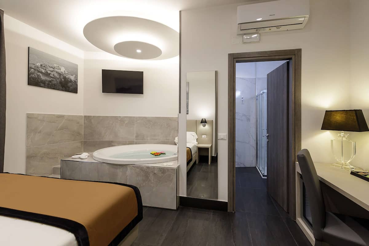 A modern hotel room with a large bed, a bathtub, a desk with a lamp, and a TV.
