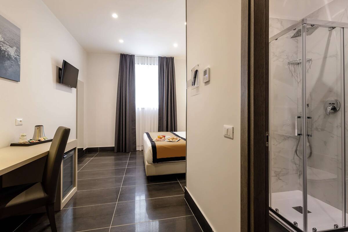 A hotel room with a bed, a desk with a chair, a TV, a shower, and a bathroom.