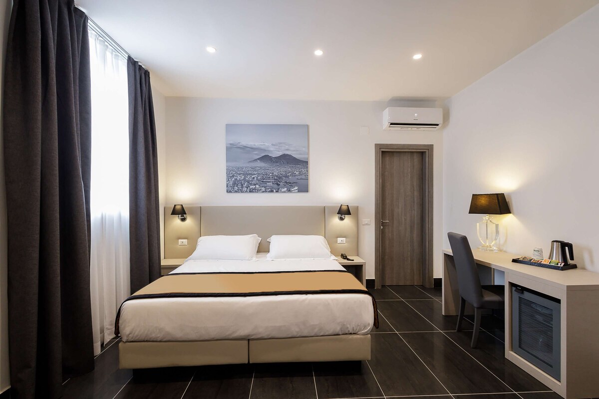 A hotel room with a bed, bedside lamps, a desk with a chair, a coffee maker, and a cityscape painting on the wall.