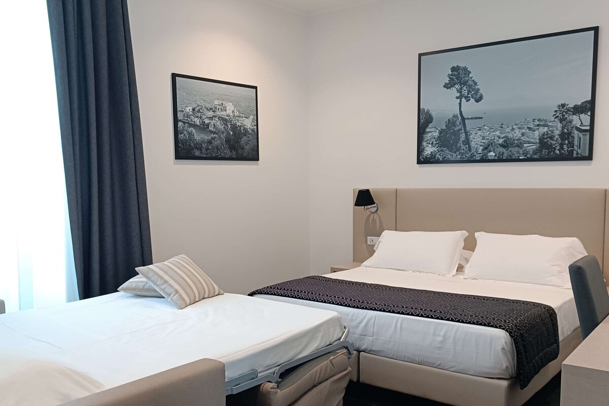 A hotel room with two beds, a large window with curtains, and two black-and-white landscape paintings on the walls.