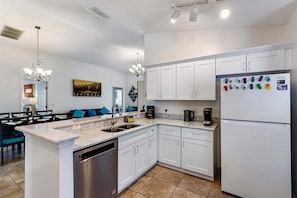 A modern kitchen with white cabinets, a dishwasher, a refrigerator with magnets, and a dining area with blue chairs.