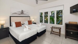 A hotel room with two beds, a ceiling fan, a large window with a view of greenery, a TV, and a small wooden table. - Beautifully appointed 1 Bedroom Ground Floor Beach Club Lagoon View Apartment (Palm Cove)