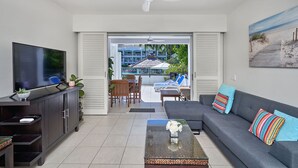 A living room with a TV, sofa, and coffee table. There is a balcony with a pool and lounge chairs. - Beautifully appointed 1 Bedroom Ground Floor Beach Club Lagoon View Apartment (Palm Cove)