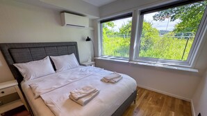 A modern bedroom with a large bed, a nightstand, a lamp, and a window with a view of greenery. - Jönköping Hotel Apartments (Jonkoping)