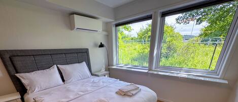 A modern bedroom with a large bed, a nightstand, a lamp, and a window with a view of greenery.