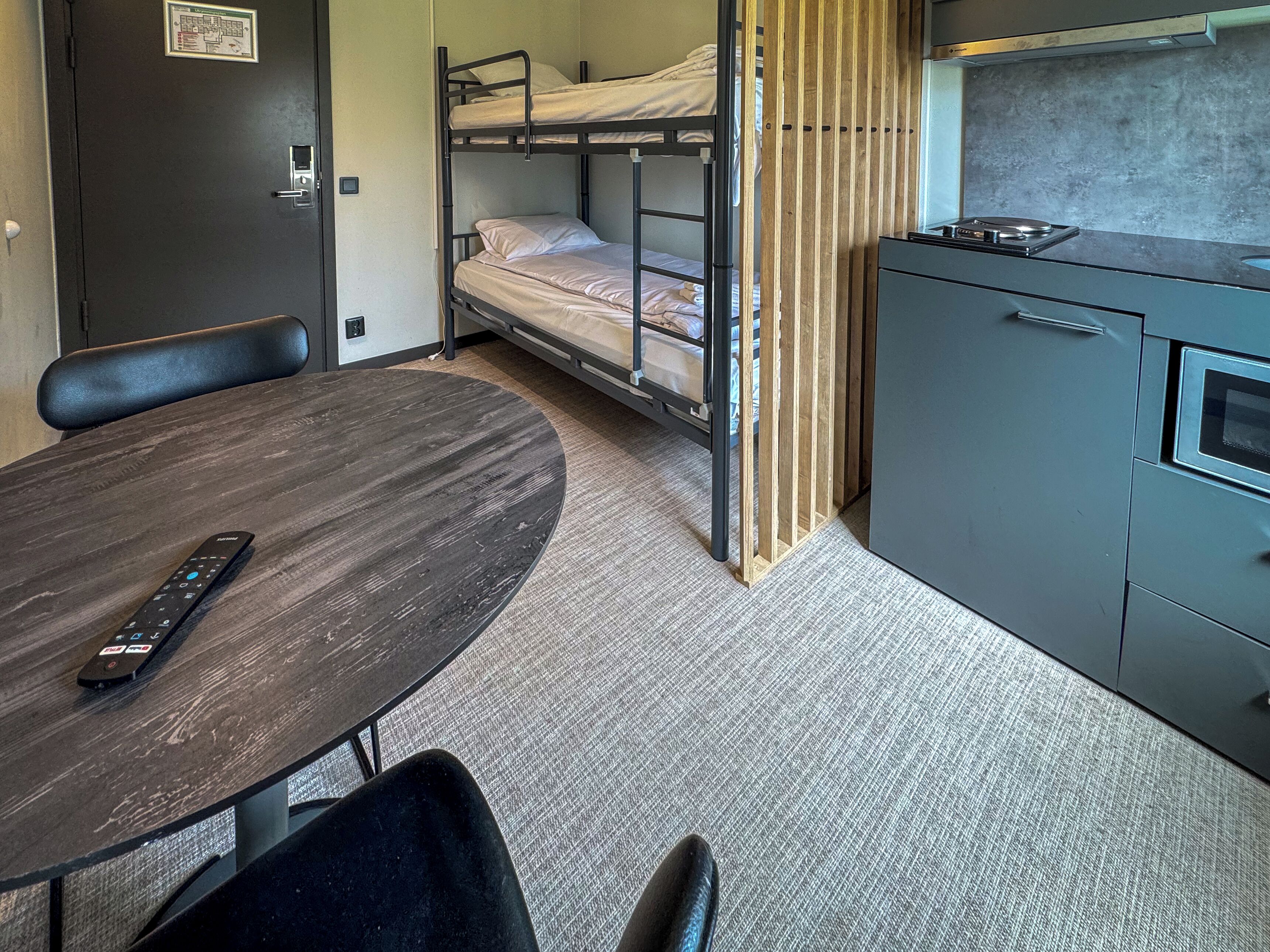 Una cama litera con separador de madera, una pequeña kitchenette, una mesa redonda con un control remoto y una silla.
