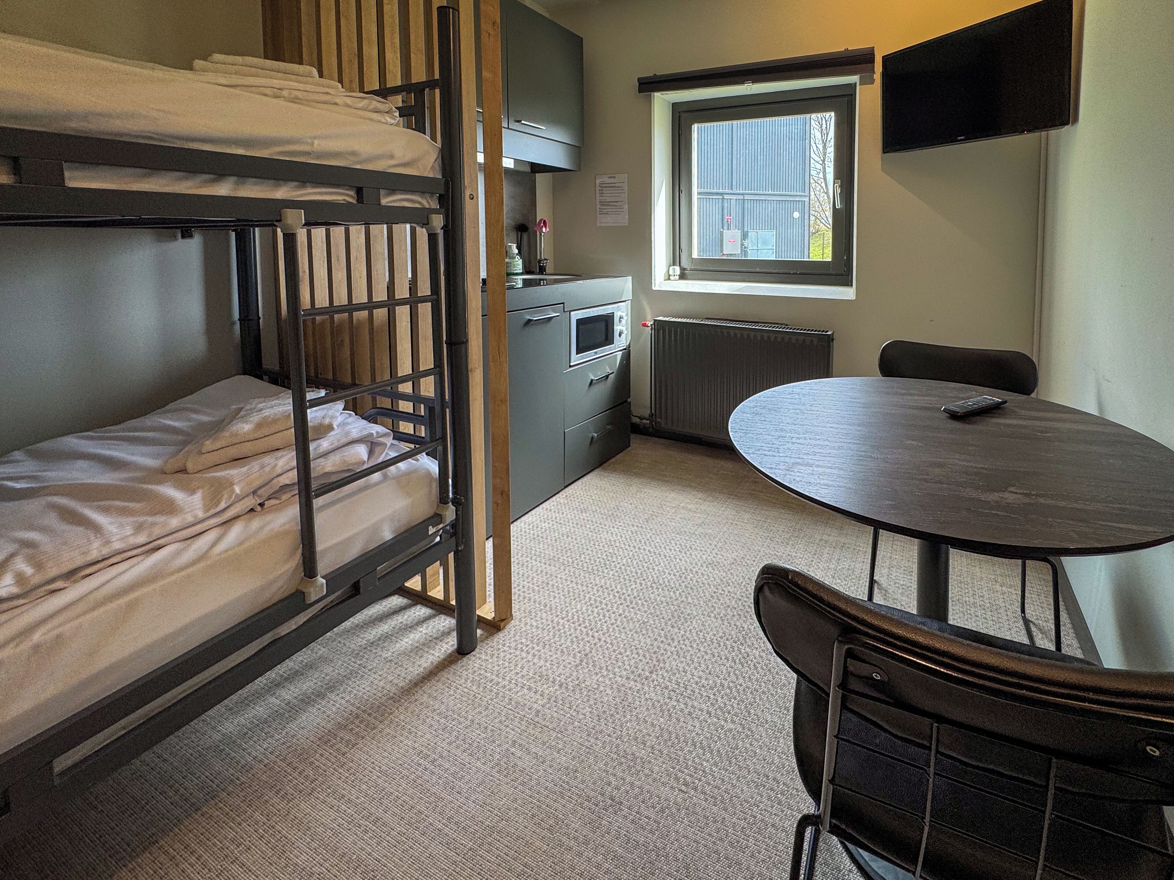 A dormitory room with bunk beds, a kitchenette, a round table, and a TV mounted on the wall.