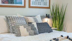 Una cama con varias almohadas, una mesita de noche con lámpara y una planta verde alta.