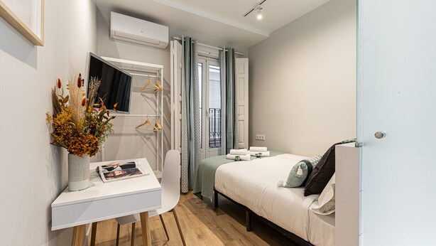 A modern bedroom with a bed, bedside table, and a small desk with a lamp and a magazine. - Waou Don Quixote Las Letras (Madrid)