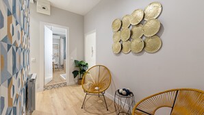A hallway with a geometric patterned wall, a modern chair, a small side table with decorative items, and a wall-mounted textured art piece. - Waou Don Quixote Las Letras (Madrid)