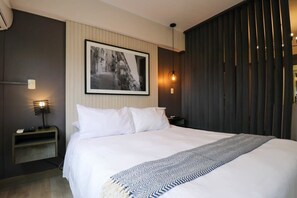 Une chambre d’hôtel moderne avec un grand lit, une photographie encadrée accrochée au mur, une table de chevet avec une lampe et un paravent sombre.