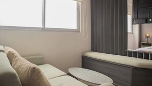 Une chambre moderne avec une grande fenêtre, un lit doté d’une tête de lit, une table de chevet et un petit tabouret.
