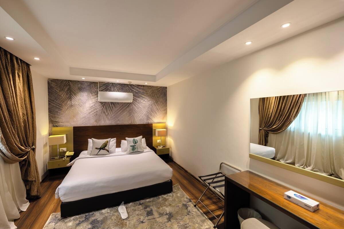 A hotel room with a large bed, bedside lamps, a mirror, and a small desk with a box.