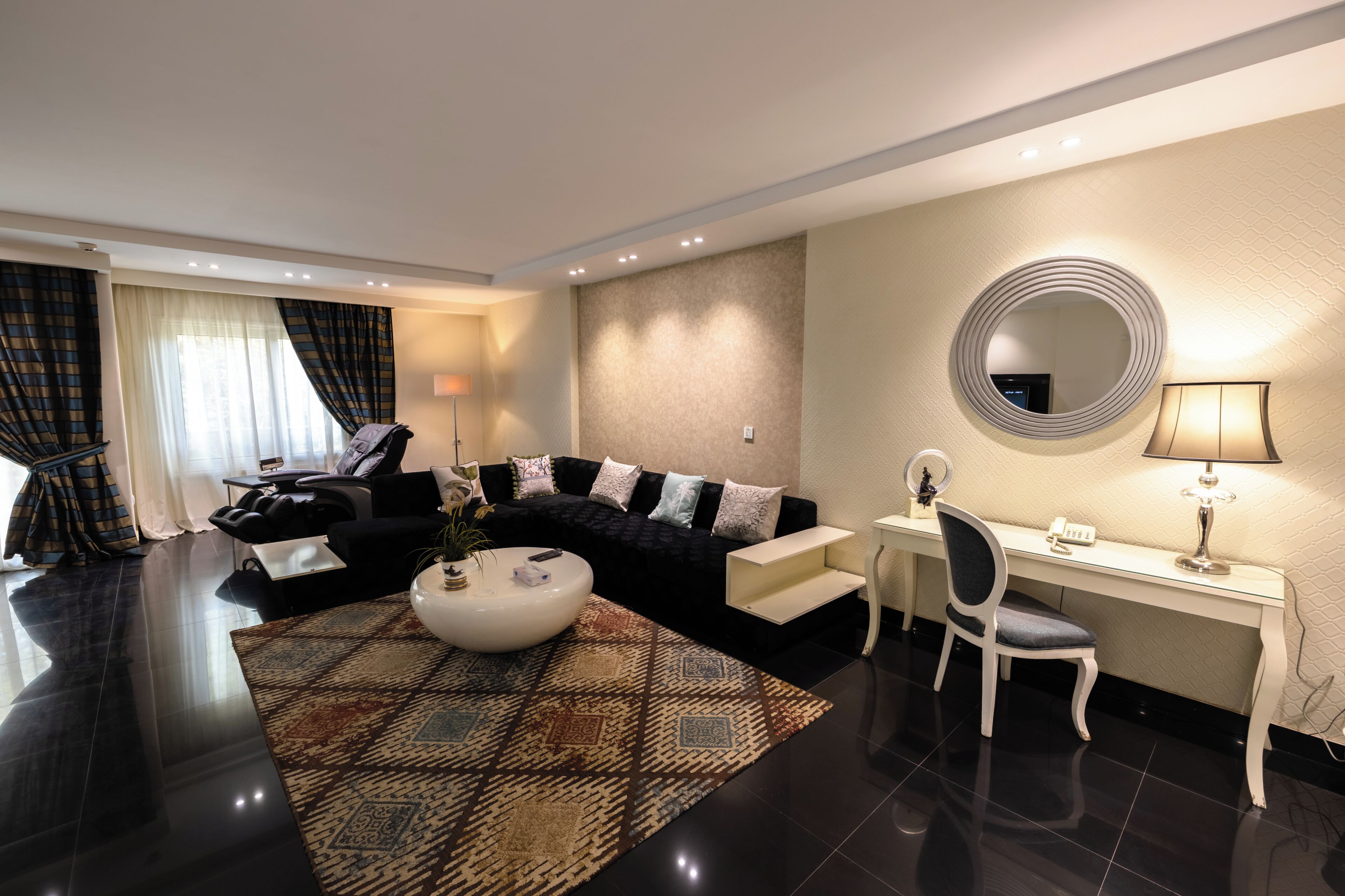 A modern hotel room with a black sofa, a white desk, a round mirror, and a patterned rug.