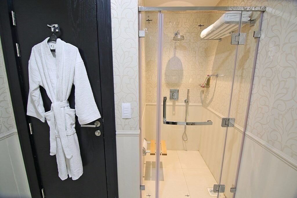 A bathroom with a glass-enclosed shower, a white robe hanging on a door, and patterned wallpaper.
