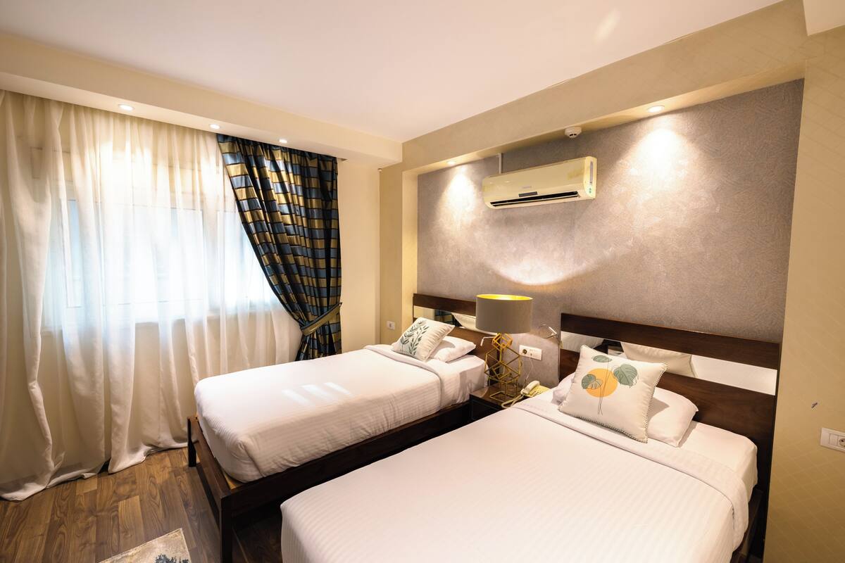 A hotel room with two beds, a wall-mounted air conditioner, and a window with sheer curtains.