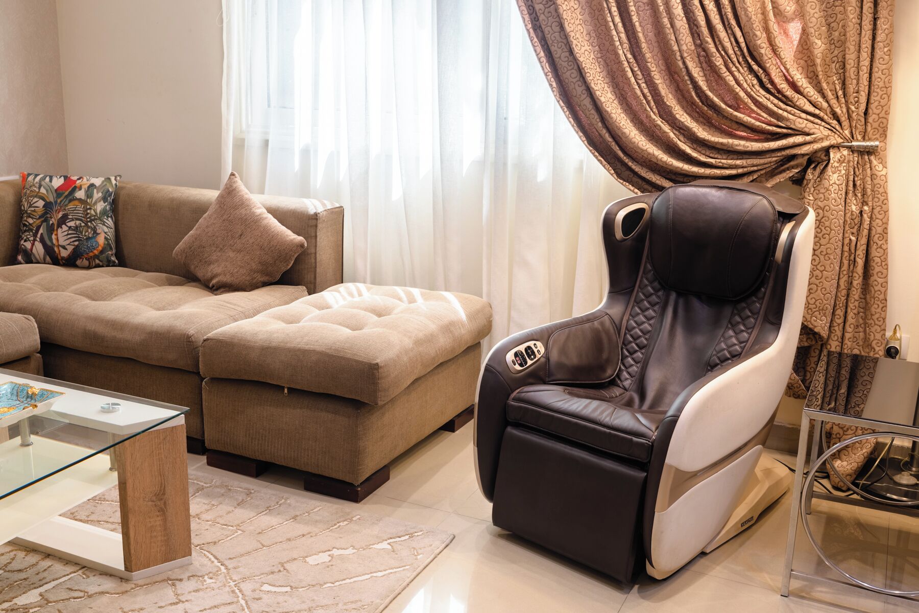 A modern living room with a sofa, ottoman, massage chair, glass coffee table, and a patterned curtain.