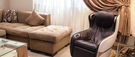 A modern living room with a sofa, ottoman, massage chair, glass coffee table, and a patterned curtain.