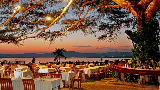 An outdoor dining area with tables and chairs under a large tree, overlooking a body of water and distant mountains. - Bagan Thande Hotel (Nyaung-U)