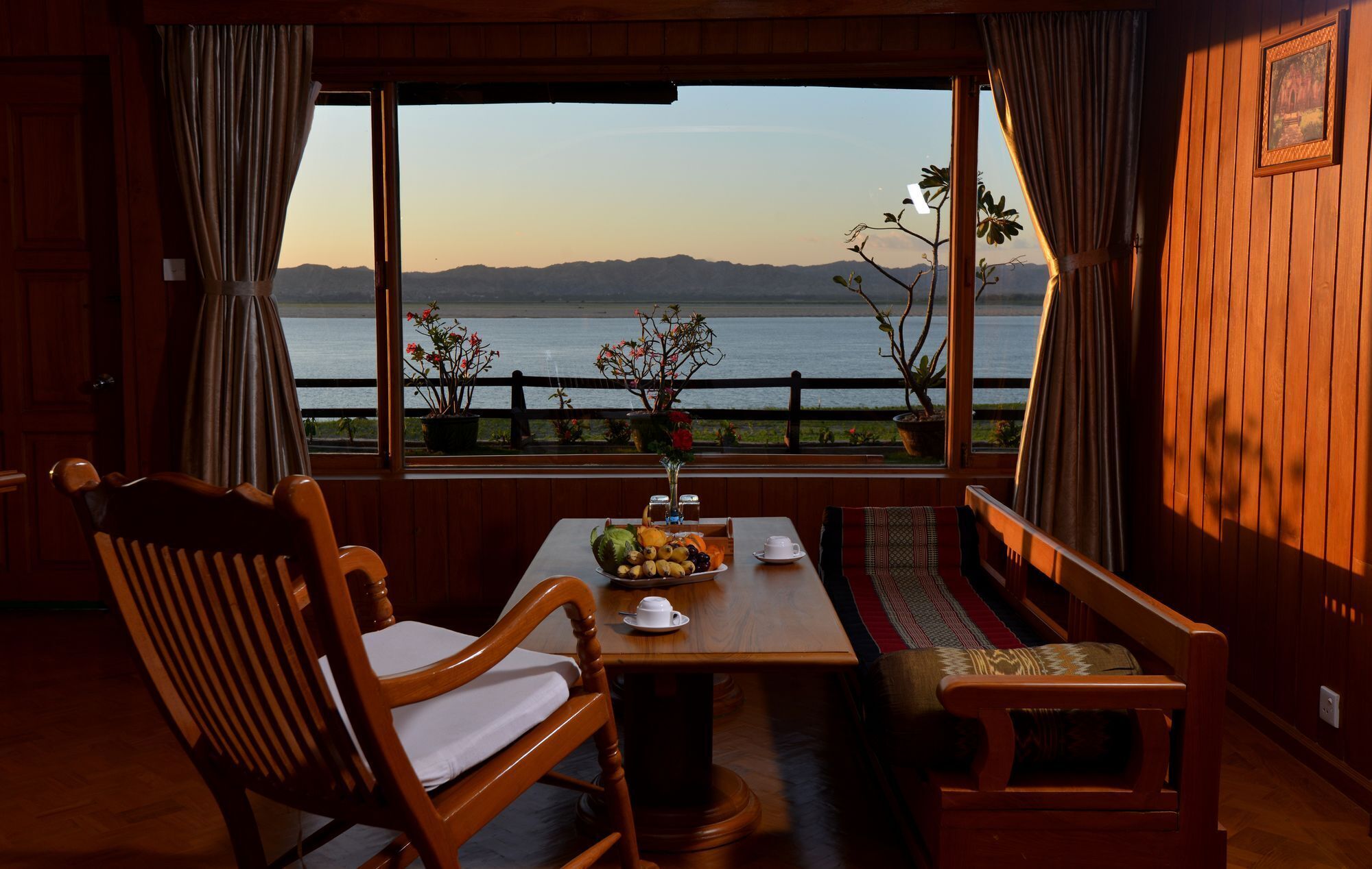 Un comedor con una mesa preparada para dos personas, sillas de madera, un banco y vistas a un lago con montañas al fondo.