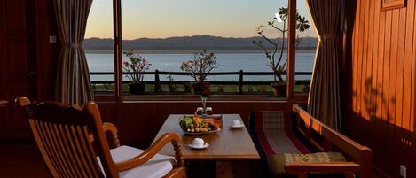 Un comedor con una mesa preparada para dos personas, sillas de madera, un banco y vistas a un lago con montañas al fondo.